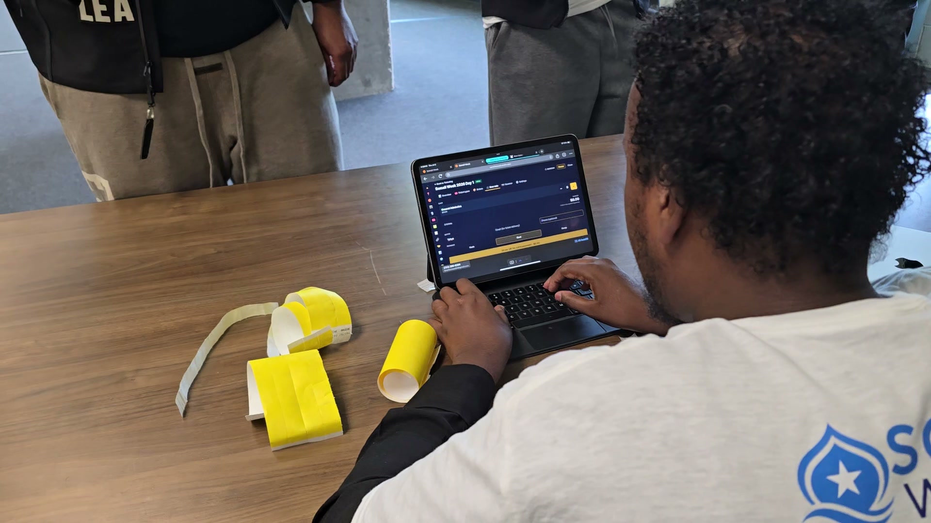 A Somali Week 2026 volunteer running door ticket sales on a laptop with the ScoreLM admin open — yellow wristbands laid out on the table next to the keyboard